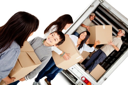 A group of five individuals in a residential area are engaged in a house removal process, standing outside near a white removals van with the rear doors open. The scene includes two women, two men, and a young boy, all actively carrying or preparing cardboard boxes of various sizes, some wrapped in packing tape. The young boy at the front, wearing a grey top, holds a medium-sized box and looks towards the camera with a smile. A woman with long dark hair, dressed in a white top and dark trousers, is handling a large box and appears to be guiding it into the van. Another woman with shoulder-length hair faces away from the camera, assisting the moving process. In the background, a man in a light-colored shirt and dark trousers is seen holding a piece of cardboard, and another man, positioned inside the van, is handing down a large box from the loading area. The scene takes place outdoors, likely on a driveway or street in Uxbridge, with a focus on the household items being carefully loaded for a house move, supported by Uxbridge Man with Van's removals service, emphasizing careful handling and organization of furniture and boxes in a typical residential setting.