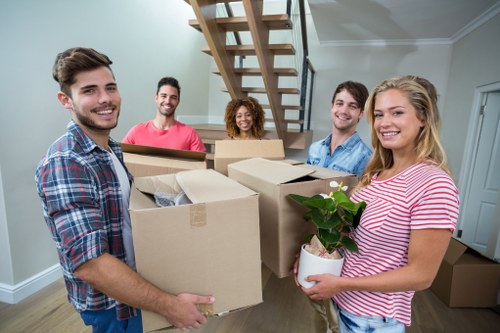 A group of five young adults, two men and three women, are smiling and standing inside a well-lit modern house during a house move, holding cardboard boxes filled with personal belongings and household items. The woman on the right, wearing a pink and white striped shirt, is holding a white pot with a green potted plant. In the background, a staircase with wooden steps and a metal railing leads to an upper floor, with an open-plan living space visible. The group is positioned in a hallway or living area with light grey walls and white ceiling moldings, surrounded by additional boxes on the floor, indicating an active move conducted by Uxbridge Man with Van, a local removals service. The scene captures a typical residential move with practical packing and careful handling of items, fitting the context of house removals in the Uxbridge area, close to Uxbridge postcode zone.