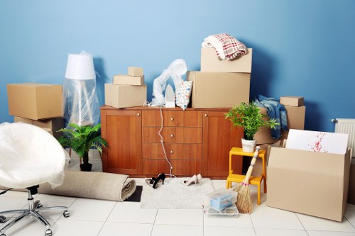 The image shows a cluttered indoor space, likely a living room in a house in Uxbridge, with a light blue wall in the background. In the foreground, there is a beige fabric sofa partially visible on the left side. The background features a wooden sideboard with multiple storage drawers, upon which various household items are placed, including several cardboard boxes of different sizes, some sealed with tape and others open, revealing packaging materials. Also on the sideboard are a plastic-wrapped lamp, a patterned cloth, and a clear bag containing packaging bubble wrap. In front of the sideboard, on the floor, there is a small white rug with a few scattered objects, including what appears to be a small black and white portable speaker, and a puppy or small dog lying on the rug. To the right, a yellow plastic step stool holds a potted plant with green foliage, and nearby are more cardboard boxes, one open showing layered items or possibly artwork. A broom with a wooden handle and natural bristles leans against the boxes. The scene is well-lit with natural or artificial lighting, suggesting an active packing or moving process in preparation for relocation within the area of Uxbridge or nearby.