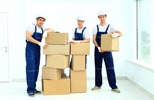 Three male removal workers in blue uniforms and white safety helmets are standing inside a bright, modern residential space with white walls and large windows, which allow natural light to illuminate the room. They are smiling and holding cardboard boxes, with two positioning boxes on top of each other, while the third holds a single box to the right. The boxes are plain brown cardboard, and the workers are wearing white gloves. The room appears to be a new or recently vacated house, with a clean, minimalistic interior and no visible furniture or personal items. The background shows the white wall and window frame, and the floor is white tiled, emphasizing the neat and professional environment. This scene exemplifies a house removal or relocation process managed by Uxbridge Man with Van, highlighting the careful handling of household items during a local move within the Uxbridge area, possibly near postcode UB8 or UB10, with the focus on efficient packing and transport of household belongings.