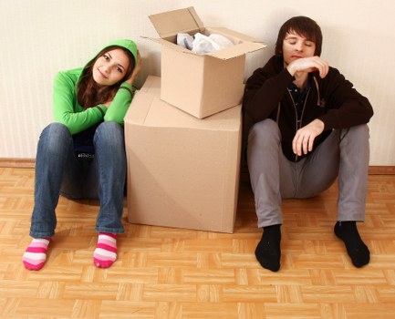 A teenage girl in a bright green hoodie and pink striped socks sits on the floor with her head resting on her arm, appearing bored or tired near a stack of cardboard boxes, with a young boy in a black hoodie and grey pants sitting beside her, also looking disengaged, in a room with a light-colored wall and wooden flooring, possibly during a house move or packing process. The boxes vary in size, some open with packing paper visible inside, and are positioned against the wall, suggesting they are ready for transportation. The scene captures a moment of pause during a household relocation, with no furniture or other household items visible, emphasizing the process of packing and moving undertaken by Uxbridge Man with Van in the local area.
