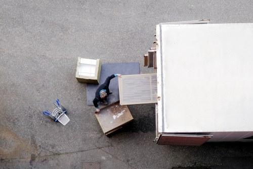 An overhead view of a professional removal worker wearing a dark jacket and a hat, inside a white removal van parked on a residential street, loading or unloading furniture and household items. The items include a small wooden chest of drawers, a grey upholstered seat or small sofa, a white wardrobe or cabinet being carried, and a cardboard box. Additional loose objects on the ground include rolled-up newspapers or magazines, a wire basket, and some plastic containers. The street surface is asphalt, and the scene is lit with natural daylight, suggesting a clear day. The overall setting indicates a house move, with the company's name, Uxbridge Man with Van, involved in residential removals in the Uxbridge area near postcode UB. The image captures the process of loading or unloading household belongings during a local relocation, emphasizing careful handling of furniture and personal items in a typical London suburb environment.
