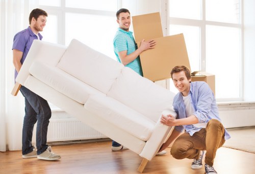 Three men are engaged in a house removal within a bright, spacious living room with large windows allowing natural light to fill the space. The man in the foreground, dressed casually in a light blue shirt and brown trousers, is kneeling and holding the arm of a white fabric sofa, which appears to be in the process of being moved. Behind him, another man in a purple shirt and jeans is lifting a large, beige upholstered armchair, supporting its frame as it is carried across the wooden floor. In the background, a third man wearing a striped turquoise T-shirt and dark trousers is holding a cardboard box, likely filled with household items or personal belongings, preparing to load it onto the sofa or move it further across the room. The scene depicts a professional and organized move, with the focus on careful handling of furniture and valuables typical of house removals services, supporting local removals operators such as Uxbridge Man with Van serving the Uxbridge area near UB8. The environment appears clean and well-lit, emphasizing a systematic approach to residential relocations.