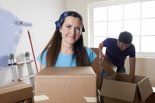 In a well-lit residential interior, a woman with long brown hair and a blue headscarf, wearing a light blue t-shirt, stands in the foreground holding a large cardboard box. The box appears sturdy, with visible tape securing its edges, and she offers a slight smile. Behind her, a young man with dark, curly hair, dressed in a dark blue t-shirt, is in the process of packing or unpacking multiple cardboard boxes of various sizes on a plain cream or light beige wall, with a large window providing natural daylight. The boxes are made of brown corrugated cardboard, some sealed with packing tape, and contain personal belongings or household items. To the left, a white wall with mounted pipework suggests the room might be an extension or part of the house undergoing relocation. The overall scene captures a typical move-in or move-out moment, emphasizing household packing activities, typical of house removal services such as those offered by Uxbridge Man with Van in the Uxbridge area, with a focus on careful handling of belongings during a residential move.
