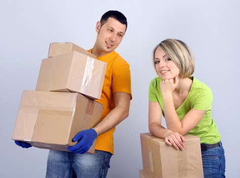 A man and a woman, both smiling, are standing indoors in a residential setting, possibly a house or apartment, preparing for a move or unloading belongings. The man is holding two cardboard boxes, one stacked on top of the other, with the top box partially taped and both appearing sturdy with visible corrugated edges. He is dressed in a bright orange T-shirt and wearing blue protective gloves, suggesting he is engaged in lifting or carrying. The woman, wearing a light green T-shirt, is leaning on a smaller cardboard box placed on a surface or the floor, with her arms resting on it and her chin supported by her hand. The background features a plain, light-colored wall, and the lighting appears natural or softly lit, creating a neutral and professional atmosphere suitable for house removals services. The cardboard boxes are of standard size with plain brown surfaces, some sealed with packing tape, implying careful packing and preparation for a residential move in Uxbridge or nearby areas. The scene conveys a collaborative and straightforward approach to home relocation within the context of professional removals.