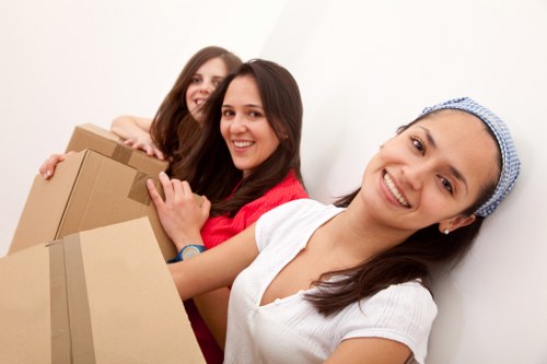 Three young women with light skin smiling and standing close together indoors against a plain white wall, each holding cardboard moving boxes. The woman in the foreground, wearing a white shirt and a blue patterned headscarf, appears to be leading the group, with her hand resting on a box. The middle woman, with long brown hair and a red top, holds a box in front of her, smiling directly at the camera. The third woman, with shoulder-length brown hair and a darker top, is positioned behind them and also smiling. The boxes are plain brown cardboard, some sealed with packing tape, and are stacked in a household environment, suggesting active packing or unpacking. The scene reflects a friendly, professional atmosphere typical of house removals services in Uxbridge or the surrounding Middlesex area, emphasizing a caring approach to relocation tasks.