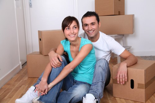 A smiling young couple, a woman with shoulder-length brown hair wearing a bright turquoise sleeveless top and blue jeans, and a man with short dark hair wearing a white t-shirt and light blue jeans, sitting on a wooden floor in a well-lit room with a white wall. They are surrounded by several large cardboard moving boxes, some stacked behind them. The boxes are plain brown with black handles, indicating they are used for a house removal. The woman is sitting cross-legged while the man sits beside her with one arm resting on a box, both facing the camera. The scene suggests they are in the process of moving into or out of a property in Uxbridge, with the focus on their happy expressions and the presence of packing equipment suitable for a professional removals service. The room has a clean, modern appearance with light wooden flooring and minimal decor, capturing an atmosphere typical of house relocation in the local area.