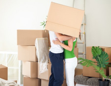 In a well-lit room with light-colored walls, two movers are engaged in a house removal in Uxbridge, with a Uxbridge postcode, as part of a professional removals service. One mover, wearing a white shirt, is lifting a large, plain cardboard box with a smooth surface and brown kraft finish, covering most of their face. The other mover, dressed in a green top and white trousers, is supporting the box from the side. Surrounding them are several other stacked cardboard boxes of various sizes, some wrapped with clear plastic, indicating packing in progress. To the right, there is a potted plant with broad green leaves placed on a small stool, adding a touch of greenery to the scene. In the background, a radiator and a door frame suggest this activity is taking place inside a residential property in Uxbridge. The overall setting depicts a typical scene of furniture and personal belongings being prepared for transport during a local house move.
