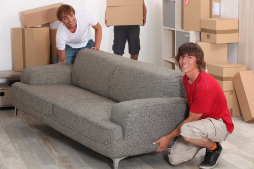 Two men are in a room with a grey fabric sofa, which they appear to be moving or positioning. One man, wearing a white shirt and jeans, is crouching behind the sofa, holding the armrest, while the other man, in a red long-sleeve shirt and beige shorts, is kneeling on one knee at the front corner, grasping the sofa's leg. The room has light-colored wooden flooring and a background of white shelving units and wall-mounted shelves filled with neatly stacked cardboard boxes, some sealed with packing tape, indicating a house removal or relocation scenario in Uxbridge. The lighting is natural or well-lit, highlighting the textures of the fabric upholstery and wooden floors. The scene reflects professional house moving services, with clear focus on furniture handling and packing within an interior space of a residential home in the Uxbridge area, supporting sustainable practices associated with local removals.
