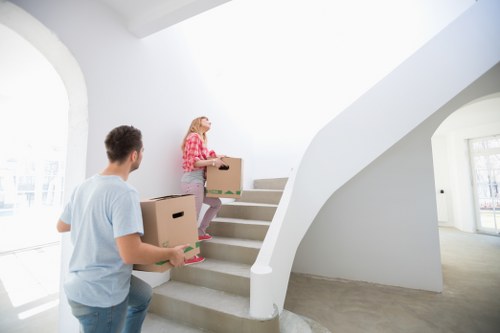 In a bright, modern home interior on a staircase, a man and a woman are engaged in a house removal activity as part of a local moving service. The man, dressed in a light blue T-shirt, is holding a medium-sized cardboard box with a pink label or tape, standing at the base of the staircase. The woman, wearing a pink and white checkered shirt and grey trousers, is on the stairs carrying a larger cardboard box, possibly containing household items or personal belongings. The staircase is white with a smooth, curved bannister, and the environment is well-lit by natural daylight coming through large arched windows visible in the background. The walls and ceiling are painted white, creating a clean and airy atmosphere, typical of a contemporary residence in Uxbridge. The scene captures the process of packing or loading items for a house move, with the staircase, boxes, and individuals actively engaged in the relocation activity, reflecting professional house removal services in the local area.