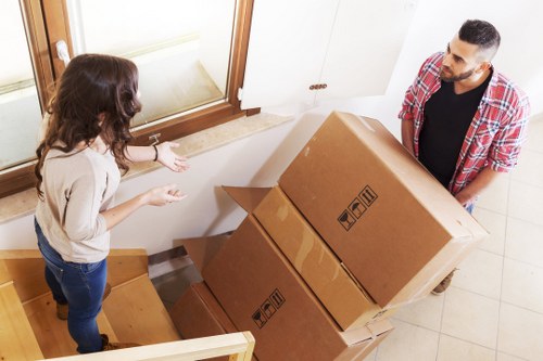 A man and a woman are inside a residential property, engaged in a conversation during a house move in Uxbridge, with the man wearing a red plaid shirt and the woman in a beige top and jeans. They are positioned near a staircase, with the woman gesturing towards a stack of three large cardboard boxes, which are sealed and have handling symbols printed on their sides. The boxes vary in size and are arranged beside a staircase leading to the upper floor of the house, which has wooden steps and a natural finish. The background shows a white wall, a window with a wooden frame allowing natural light into the space, and a beige tiled floor. The scene captures the process of loading or unloading household items in a typical home setting during a professional removals service provided by Uxbridge Man with Van, emphasizing safe handling and organization during a house relocation.