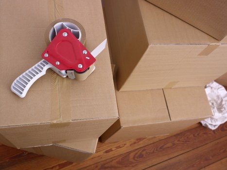 A close-up view of a cardboard moving box on a polished wooden floor in a residential setting, with a red and white tape dispenser placed on top of the box. The box is sealed with brown packing tape and appears to be part of a house removal process in Uxbridge, with nearby additional boxes stacked in the background. The scene is well-lit, highlighting the texture of the cardboard and the glossy finish of the tape dispenser, indicating a professional house removals service by Uxbridge Man with Van preparing items for relocation within the local area. The focus is on the packing materials, suggesting the process of packing or sealing belongings for a move from a household in the town, with the environment indicating an interior residential space.