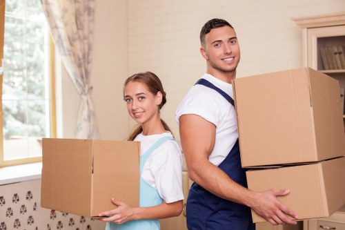 A young man and a young woman are inside a well-lit room, possibly a living room or hallway, with cream-colored walls and a window decorated with floral curtains in the background. The man is smiling and holding two medium-sized cardboard boxes, one stacked on top of the other, with a natural brown finish. He has short, dark hair and is wearing a white t-shirt with navy blue overalls. The woman, standing slightly behind and to the left of him, is also smiling while holding a smaller cardboard box, with her hair tied back and wearing a white shirt under a light blue apron. The boxes appear to be empty or filled with household items, typical of house removals or relocation preparations. The scene suggests an organized moving process with both individuals actively participating in packing or transporting belongings, with the environment indicating a residential setting in Uxbridge or nearby town, consistent with the local house removals services provided by Uxbridge Man with Van.