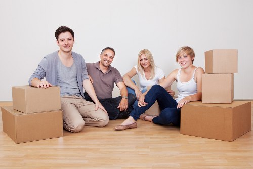 Four young adults, two men and two women, sit on a light wooden floor in a spacious, well-lit room with plain white walls. They are surrounded by several cardboard boxes, some stacked to the right and left, indicating a house moving or packing process, typical of a Uxbridge-based removals service. The individuals appear relaxed and happy, with casual clothing—one man wears a gray jacket over a T-shirt, another in a polo shirt, the women in light-colored tops and jeans. The boxes are plain brown with no visible labels, and their positions suggest they are in the midst of packing or unpacking. The room has no furniture or other visible household items, focusing attention on the boxes and the group. The overall scene captures a typical moment during a professional house move within the Uxbridge area, aligning with the company's service category of removals and emphasizing a friendly, organized moving process.