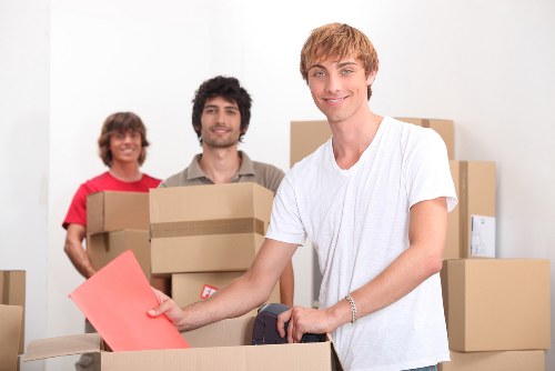 A young man with light brown hair and fair skin, wearing a white T-shirt, is smiling and holding a pink envelope or document while standing amidst open cardboard moving boxes in a bright, indoor residential space. Behind him, two other individuals, a woman with curly short brown hair wearing a red top and a man with dark, slightly wavy hair wearing a beige shirt, are also smiling and holding additional cardboard boxes, indicating a house removal or relocation process. The setting features a plain white wall with no visible furniture, emphasizing the focus on the packing activity. The scene suggests a professional house removals service in the Uxbridge area, with the team engaged in organizing and transporting household items, ready to assist with moving homes within or near Uxbridge, UB8 postcode. The lighting is natural and even, highlighting the collaborative effort involved in a local move handled by Uxbridge Man with Van.