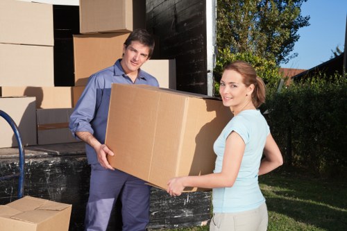 A man and a woman, both appearing to be in their late twenties or early thirties, are standing outdoors next to an open removals van on a residential street near Uxbridge, Middlesex, during daytime. The man, dressed in a blue shirt and dark trousers, is holding a large cardboard box and smiling at the camera. The woman, wearing a light blue T-shirt and beige trousers, is also holding the box, with her body slightly turned to the side and smiling. In the background, the side of the black removals van is open, revealing several stacked cardboard boxes of various sizes, some with visible tape, ready for relocation. The area around them is green with a well-maintained lawn, bushes, and trees, with a small house or shed visible in the distance. Natural daylight illuminates the scene, casting soft shadows and highlighting the textures of the cardboard, clothing, and greenery, reflecting the typical household items involved in residential removals and demonstrating professional moving assistance provided by Uxbridge Man with Van in the local Middlesex area.