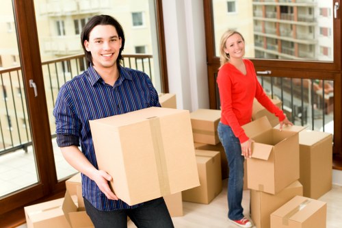 A young man and woman are inside a well-lit, modern apartment with large windows and a view of a residential street, likely in Uxbridge or nearby in the UB postcode area. The man, positioned on the left, is holding a medium-sized cardboard box securely with both hands, and he is smiling directly at the camera. He has dark hair, light skin, and is dressed in a striped blue and black shirt. The woman, on the right, is standing next to a stack of brown cardboard boxes on the floor and is also smiling while holding an open box, appearing to be in the process of packing or unpacking. She has blonde hair, light skin, and is wearing a bright red long-sleeved top with blue jeans. The room's interior features wooden window frames and a balcony outside the window. The scene is bustling with activity typical of house removals, with generic cardboard boxes of various sizes stacked around, some sealed with packing tape. This image illustrates a professional house removals service, likely provided by Uxbridge Man with Van, capturing a moment of packing at a residence in or near Uxbridge. The natural lighting highlights the clean, organized space and emphasizes the personal effort involved in a house move, complementing the company’s local removals services.