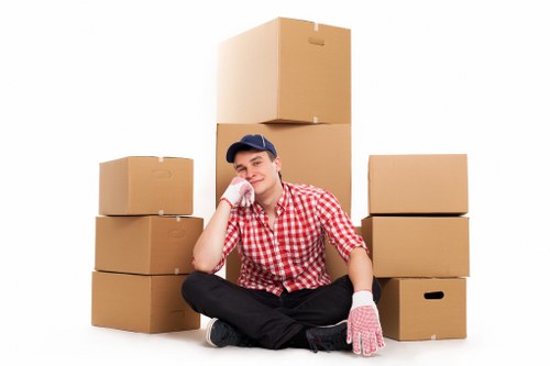 A young man dressed in a red and white checkered shirt, wearing a navy baseball cap and pink work gloves, sitting cross-legged on the floor in front of a stack of six cardboard moving boxes. The boxes vary in size and are neatly arranged, with three on each side of the man. The background is plain white, highlighting the focus on the individual and the boxes, which suggest a house removal or packing activity. The man appears relaxed, with a slight smile, and is resting his chin on his right hand. His facial expression and posture indicate a moment of pause during a house relocation process. The scene emphasizes organized packing, typical of professional removals services like those offered by Uxbridge Man with Van in the Uxbridge area, offering a visual representation of ongoing household packing tasks associated with residential moves in the local area, including nearby postal code regions in Uxbridge, Middlesex.