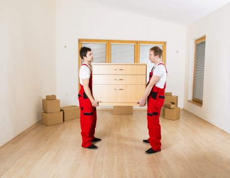 Two movers dressed in white shirts and red trousers with black knee pads are positioned face-to-face in a bright, empty room, each holding a large wooden chest of drawers with a light finish and multiple drawers. The room has white walls, a wooden floor, and a large window with closed blinds on the right side, allowing natural light to illuminate the scene. In the background, a double-glazed window with a wooden frame is visible, along with several stacked cardboard boxes placed on the floor near the walls, indicating a house move or relocation process. The movers are carefully balancing the furniture piece between them, emphasizing careful handling typical of professional removals. The environment appears interior, residential, and is part of a house in Uxbridge or surrounding area, as suggested by the context of Uxbridge Man with Van services. The scene captures the careful, methodical approach involved in house removals, highlighting the furniture handling aspect central to such services, and subtly referencing local house moving activities.