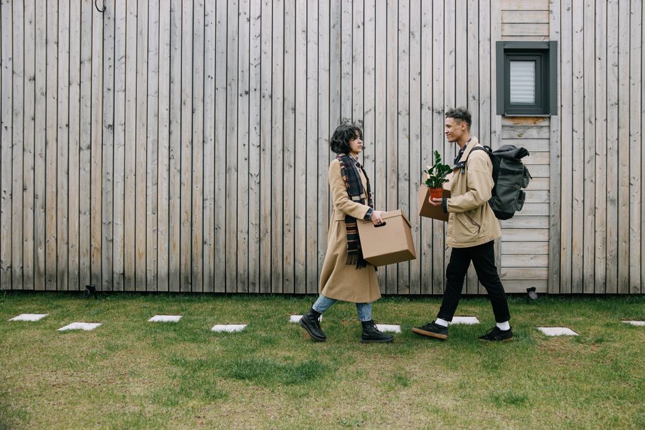 A young woman and man sit on a staircase inside a building, each holding a cardboard box as part of a house relocation process. The woman, dressed in a checked shirt, white top, and sneakers, holds a 